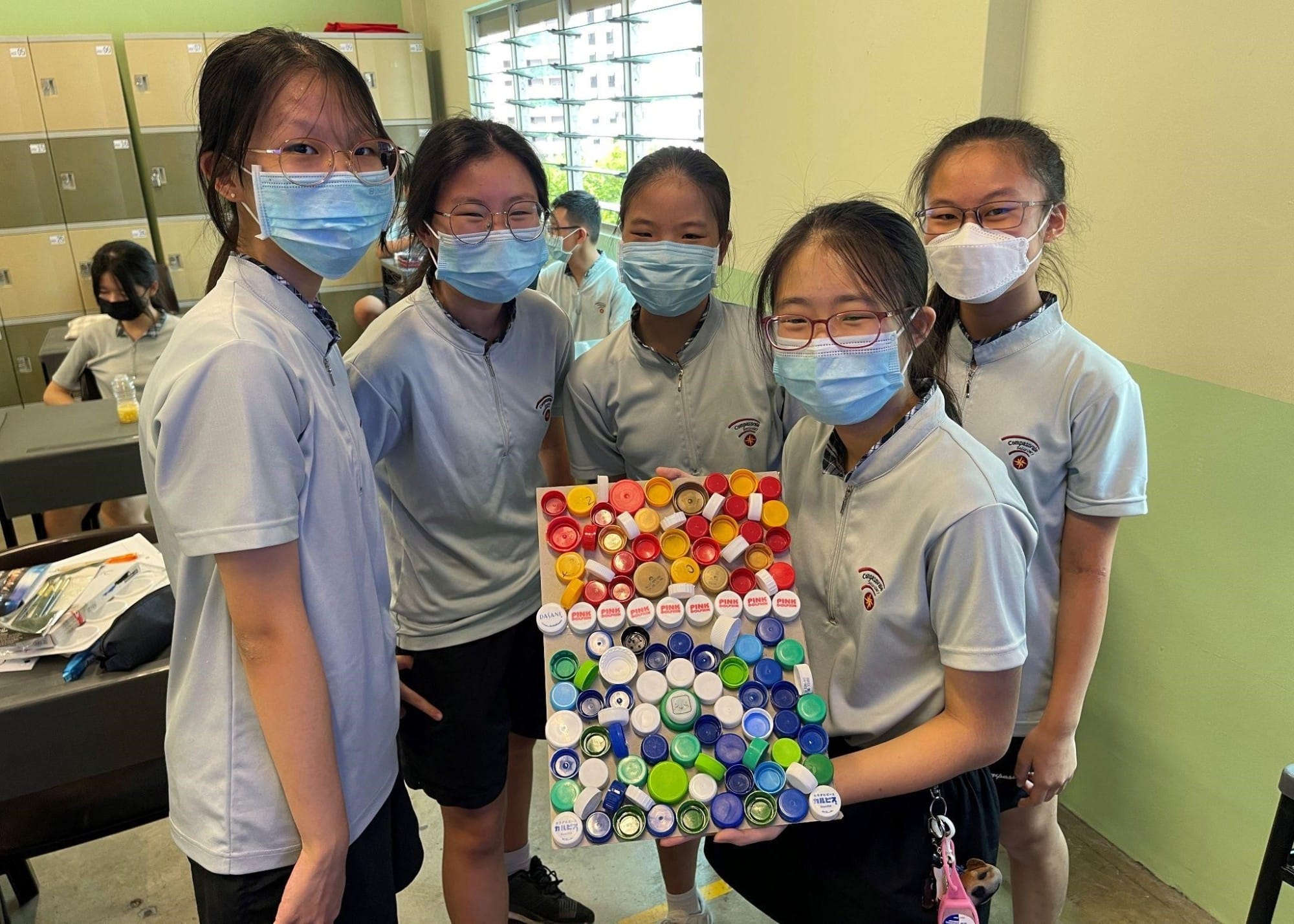 Five masked students in school uniforms hold a board covered with colorful plastic bottle caps arranged in a pattern.